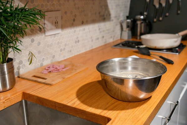Bancada de madeira com tigela de inox contendo massa de preparo, tábua com presunto picado, planta decorativa e fogão ao fundo, ilustrando a rotina de organização durante o preparo de refeições.