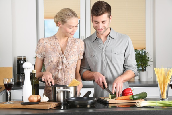 Casal cozinhando junto em uma bancada moderna; o homem corta legumes em uma tábua de madeira enquanto a mulher mexe o macarrão em uma panela de inox.