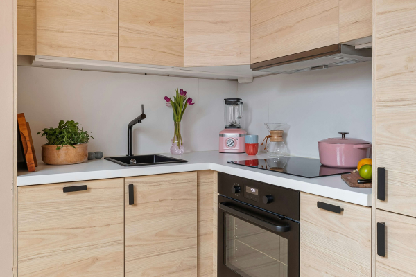 Cozinha moderna com armários de madeira clara e bancada branca; sobre a bancada, um liquidificador rosa, vaso com tulipas roxas, bule de café de vidro e uma panela rosa sobre o cooktop de indução