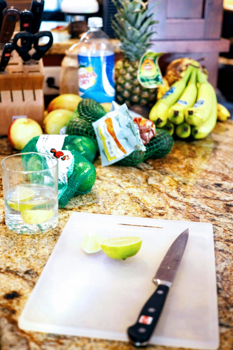 Bancada de granito com frutas frescas como abacaxi e bananas ao fundo, e uma tábua de corte com limões fatiados e faca em primeiro plano, ilustrando a organização de alimentos.