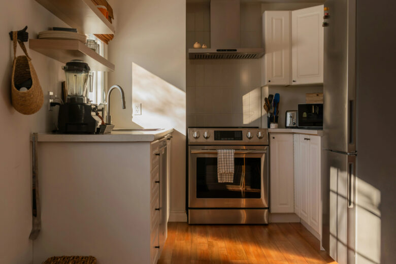 Cozinha moderna com eletrodomésticos de inox, bancada branca com liquidificador e cafeteira, e piso de madeira iluminado por uma forte luz solar diagonal.