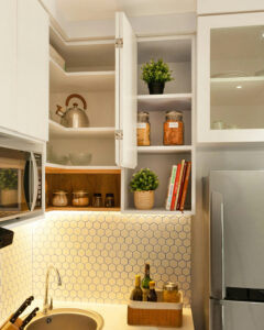 Armário de canto de cozinha branco aberto com prateleiras organizadas contendo livros de receitas, chaleira de inox, potes de vidro e plantas decorativas sob iluminação LED.