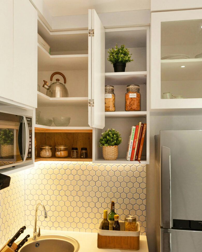 Armário de canto de cozinha branco aberto com prateleiras organizadas contendo livros de receitas, chaleira de inox, potes de vidro e plantas decorativas sob iluminação LED.