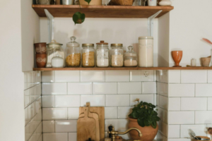 Prateleira de madeira instalada em parede de azulejos brancos com potes de vidro organizados, tábuas de carne e planta decorativa em uma cozinha rústica e funcional.