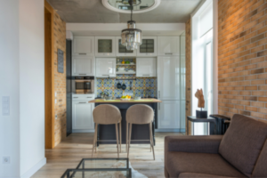 Cozinha integrada moderna com bancada escura, banquetas bege, armários brancos e backsplash de azulejos coloridos, vista a partir de uma sala com sofá marrom e tapete felpudo.