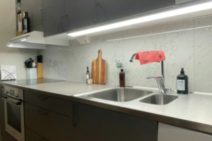 ozinha moderna com fita de LED instalada sob os armários superiores, iluminando a bancada de inox, a pia e o fogão de indução em um ambiente pequeno e funcional.