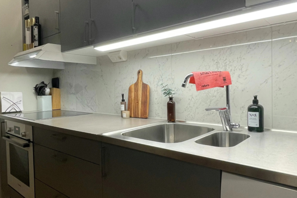ozinha moderna com fita de LED instalada sob os armários superiores, iluminando a bancada de inox, a pia e o fogão de indução em um ambiente pequeno e funcional.